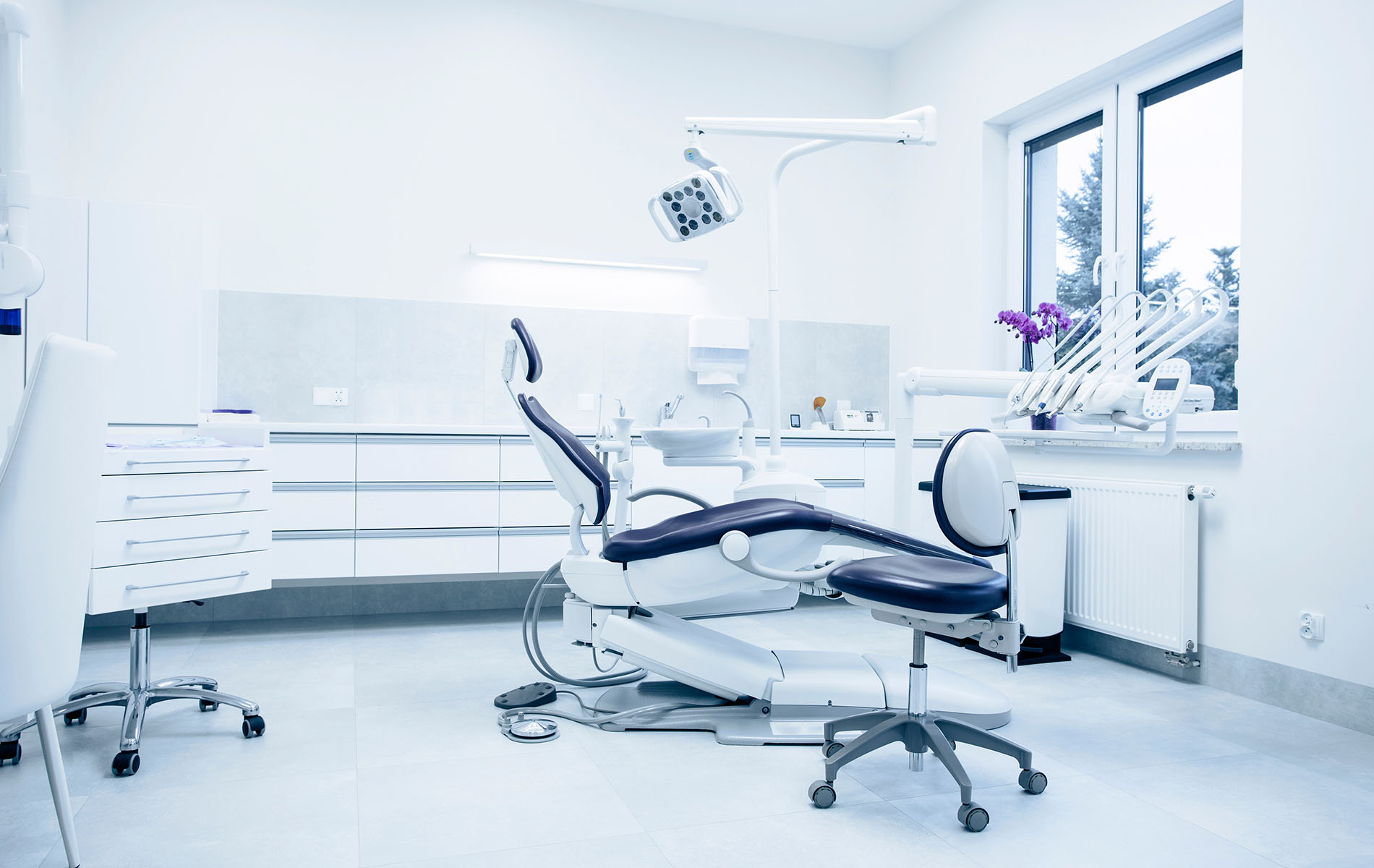 The image shows a modern dental office interior with a dental chair and equipment, surrounded by white walls and cabinets, under bright lighting.